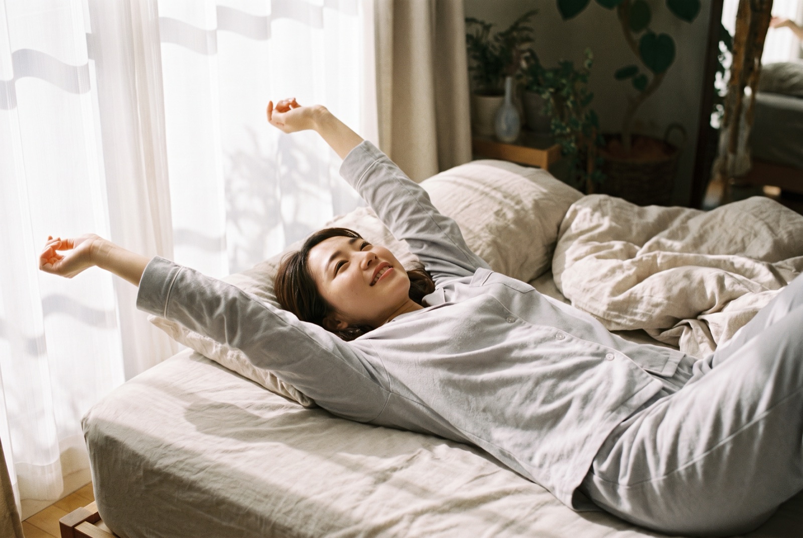 布団の中で仰向けのまま、両手を頭の上に伸ばして大きく伸びをしている女性