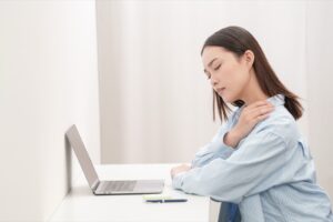 パソコンの前で、首の後ろに手を当てて顔をしかめる女性