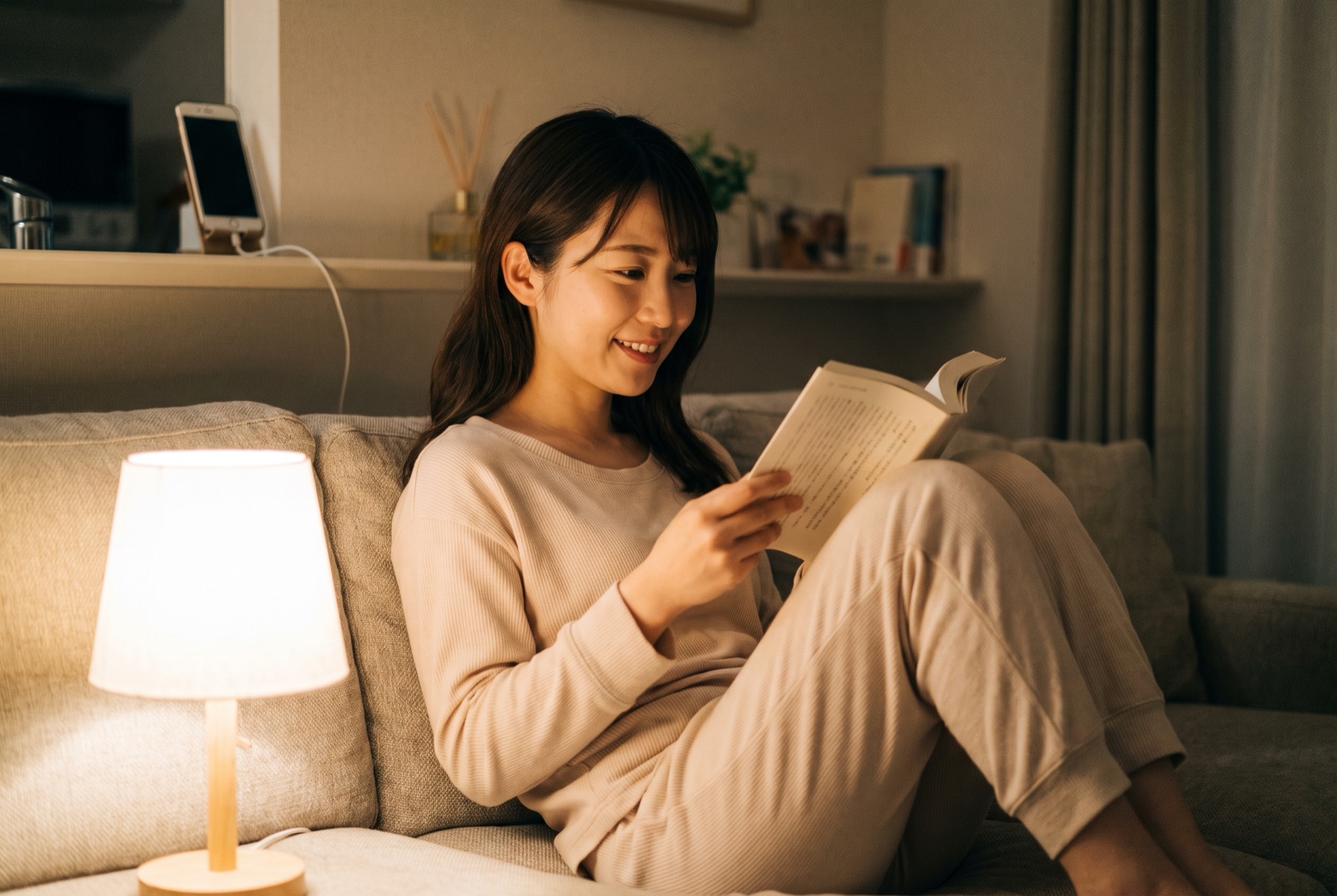 天井の明かりを消し、スタンドの明かりだけの部屋で紙の本を読む女性。スマートフォンは離れた棚で充電している
