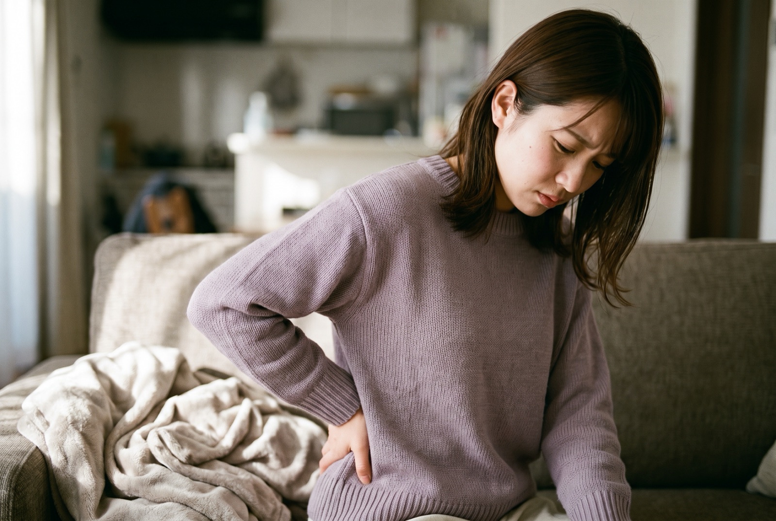 ソファのそばで、片手で腰の後ろを押さえてつらそうにしている女性