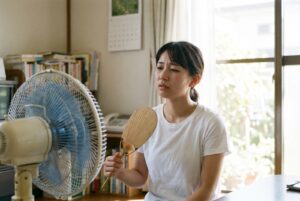 夏の終わりの部屋で、扇風機の前に座り、うちわを持ってぐったりしている女性